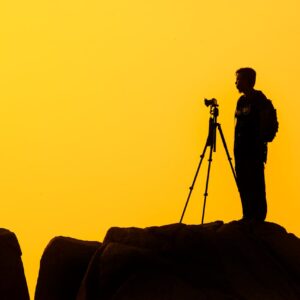 Silhouette of a lone photographer with tripod on a rocky cliff against a golden sunset sky.